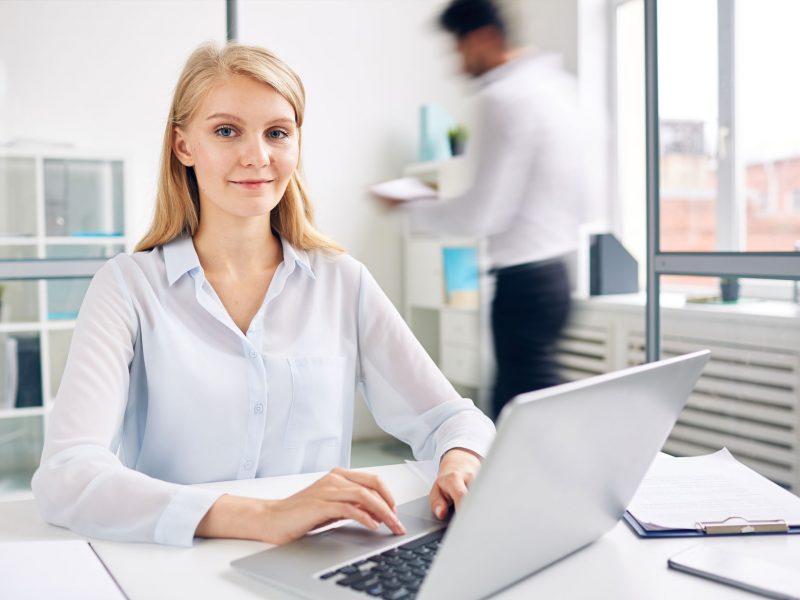 Successful young businesswoman with laptop typing by workplace in office environment